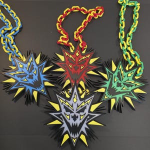 May include: A collection of colorful necklaces featuring starburst pendants with menacing faces. The pendants are in blue, red, green, and gray, with yellow accents. The chains are in matching colors, creating a bold and eye-catching design.