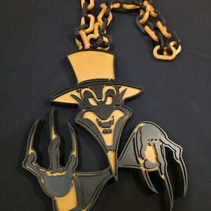 Pode incluir: Um pendente preto e dourado que retrata uma figura de diabo com cartola e mãos com garras, suspenso por uma corrente a condizer. O design tem um efeito 3D em camadas. O tema do pendente é gótico ou inspirado no Halloween.