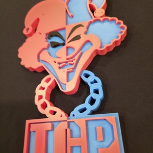 Puede incluir: Una cara de payaso bicolor, roja y azul, impresa en 3D, con las letras "ICP" en la parte inferior. El payaso tiene una sonrisa siniestra y está rodeado por un contorno azul. Las letras también son rojas y azules.