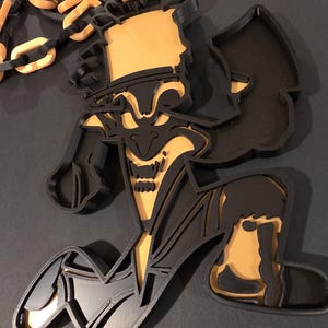 Op de afbeelding: Een gelaagde 3D-geprinte wanddecoratie met een gestileerd personage met een hoge hoed, uitgevoerd in matzwart en goudkleurig plastic. De figuur staat in een dynamische, gehurkte houding met een ketting van afwisselend zwarte en gouden schakels.