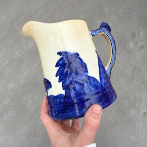 Könnte beinhalten: Eine handgefertigte Keramikkrug mit cremefarbenem Körper und blauem Blumendesign. Der Griff und der Rand sind ebenfalls blau. Der Krug wird von einer Hand gehalten.