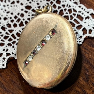 May include: A round gold-tone metal locket featuring a diagonal bar set with alternating clear and deep red stones. The locket has a smooth, slightly worn surface and includes a small circular bail at the top for attachment to a chain or cord.
