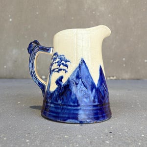 Könnte beinhalten: Eine Keramikkrug mit cremefarbenem Oberteil und blauem Berglandschaftsdesign. Der Griff und der Boden sind blau. Der Krug hat einen Ausguss zum Ausgießen. Das Design beinhaltet einen Baum und eine Figur.
