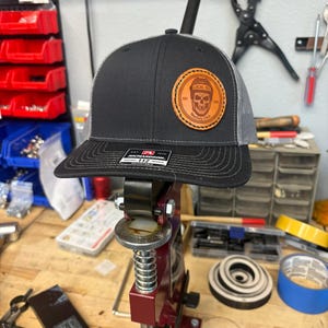 Könnte beinhalten: Eine schwarz-graue Trucker-Cap mit einem runden, hellbraunen Lederaufnäher auf der Vorderseite. Der Aufnäher zeigt einen Totenkopf mit Eishockeyhelm und den Text "LOCAL 192" sowie "Hockey Club". Ein schwarzer Aufkleber auf dem Schirm zeigt "RICHARDSON 112 ADJUSTABLE".