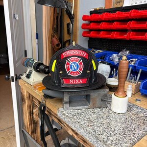 May include: A black leather firefighter helmet front piece featuring a central red and white Maltese cross emblem with the letters F, N, and D. The piece includes red banners with the white text JOHNSON and NIXA, and MO, with yellow accents.