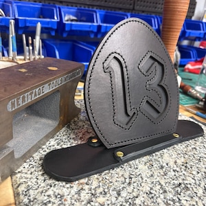 Puede incluir: Una pieza frontal de cuero negro para casco de bombero con el número 13 grabado en el centro. El escudo está cosido con hilo negro y sujeto a una base de cuero horizontal con dos remaches metálicos de color latón en la parte inferior.