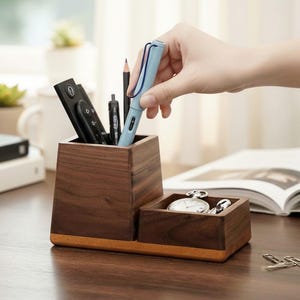 May include: A wooden desk organizer with two compartments. One holds pens, a remote, and a pencil. The other holds a pocket watch. A light blue pen is being placed in the organizer. The organizer is on a wooden desk.