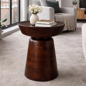 Może przedstawiać: Ciemnobrązowy drewniany stolik boczny z okrągłym blatem i podstawą w kształcie cokołu. Na stole znajduje się biały wazon z kwiatami i książkami. Stół znajduje się w salonie.