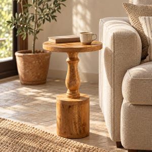 May include: A round wooden side table with a smooth top, a central turned pedestal column, and a cylindrical base. The table is crafted from light brown wood with a visible grain pattern. A small book and a ceramic mug sit on the table surface.