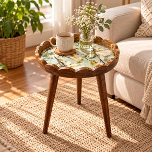 Peut inclure: Une table d'appoint ronde en bois avec trois pieds fuselés et un bord festonné. Le plateau présente une surface vert clair avec un motif floral de magnolia imprimé. Une tasse en céramique blanche et un vase en verre avec des fleurs blanches.