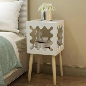 Pode incluir: Uma mesa de cabeceira branca com um design de recorte ondulado único nas laterais. A mesa tem quatro pernas de madeira e uma prateleira com livros e uma chávena de chá. Um vaso prateado com flores brancas está em cima.