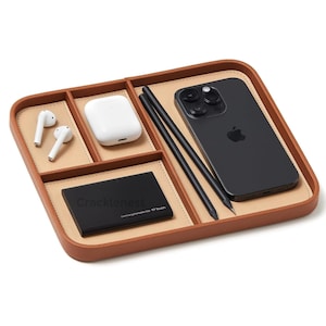 May include: A brown leather desk organizer with multiple compartments. It contains a black smartphone, two pencils, a white charging case, wireless earbuds, and a black external hard drive. The organizer has a light tan interior.