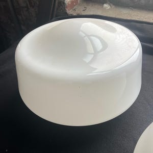 May include: A white, opaque, cylindrical lampshade with a rounded top. The lampshade has a smooth, glossy surface and reflects light. The lampshade is likely made of glass or a similar material. The overall shape is a simple, classic design.