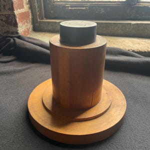 May include: A wooden object with a dark gray top, a cylindrical middle section, and a tiered base. The wood has a warm, natural tone. The object is likely a decorative item or a small stand.