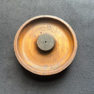 May include: A round, wooden ashtray with a central, raised, dark gray circle. The ashtray is made of light brown wood with visible grain patterns. The ashtray has a shallow bowl shape with a slightly raised rim.