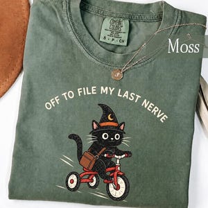 May include: A moss green t-shirt featuring a graphic of a black cat wearing a witch hat and riding a red tricycle. The cat carries a brown satchel. The text "OFF TO FILE MY LAST NERVE" is printed in white above the cat. A gold necklace rests on the shirt.