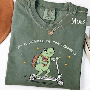 Puede incluir: Una camiseta color verde musgo con el gráfico de una rana verde claro que lleva una mochila marrón y monta un patinete blanco. La rana sostiene dos lápices de colores. El texto dice "OFF TO WRANGLE THE TINY TORNADOES" con pequeñas estrellas.