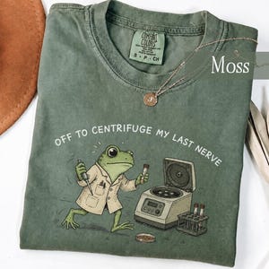 May include: A moss green t-shirt featuring a graphic of a frog wearing a lab coat, holding a pipette, and standing next to a centrifuge. The text reads, "OFF TO CENTRIFUGE MY LAST NERVE." The shirt has a crew neckline and a Comfort Colors brand label.
