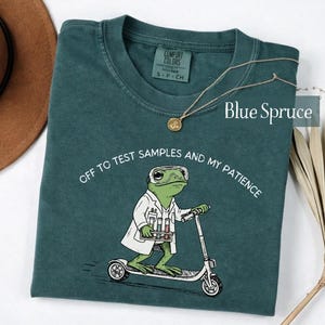 May include: A dark teal t-shirt featuring a graphic of a green frog wearing a white lab coat and safety goggles while riding a scooter and holding a test tube rack. The text reads, "OFF TO TEST SAMPLES AND MY PATIENCE." The shirt has a Comfort Colors label.