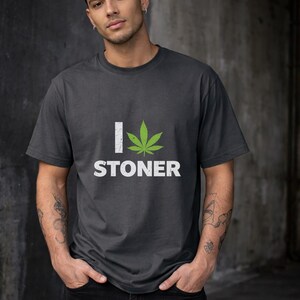 May include: A dark grey t-shirt featuring the text "I Stoner" with a green cannabis leaf graphic. The shirt is made of a soft material and is suitable for casual wear. The design is printed in white.