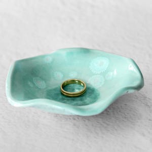May include: A small, shallow, wavy-edged ceramic dish in a light mint green glaze with white crystalline speckles. A simple, smooth gold-toned metal band ring rests in the center of the dish, which features a darker green circular base.