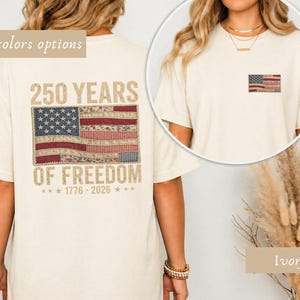 Puede incluir: Camiseta de color marfil con un diseño de bandera estadounidense desgastada. La parte posterior de la camiseta muestra un gráfico de bandera más grande con el texto "250 YEARS OF FREEDOM 1776-2026". La camiseta tiene cuello redondo y mangas cortas.