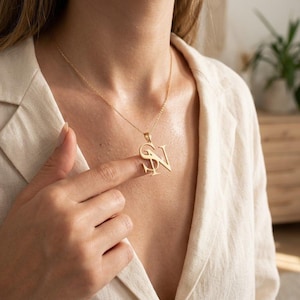 Puede incluir: Un collar de tono dorado con un colgante que presenta las letras mayúsculas S y N entrelazadas. El colgante cuelga de una cadena fina y delicada de tono dorado. Las letras están conectadas en un diseño estilizado y superpuesto con un acabado pulido.