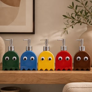 Könnte beinhalten: Fünf farbenfrohe Keramik-Seifenspender in Form von Pac-Man-Geistern. Jeder Spender hat eine silberne Pumpe und Wackelaugen. Die Farben sind grün, blau, gelb, rot und braun, angeordnet auf einem Holzregal.
