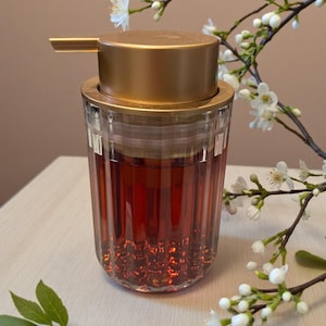 May include: A clear glass soap dispenser with a gold-coloured pump and lid. The dispenser is filled with a reddish liquid. White flowers and green leaves are in the background, creating a natural aesthetic.