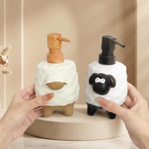 May include: Two ceramic soap dispensers. One is white with a brown wooden pump and a brown oval shape. The other is white with a black pump and a black sheep face design. Both have brown feet.