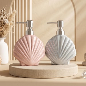 May include: Two ceramic soap dispensers in the shape of seashells, one pink and one grey, with silver pump heads. They sit on a beige platform. The background is a neutral colour scheme with decorative items.