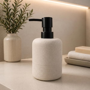 Op de afbeelding: Een cilindrische zeepdispenser met een matte, gebroken witte, gespikkelde behuizing en een zwart pompmechanisme. Het onderste gedeelte van de dispenser is voorzien van een reeks horizontale ribbels, terwijl het bovenste deel glad is.