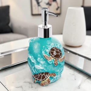 May include: A teal ceramic soap dispenser with a chrome pump. The dispenser features two brown and tan sea turtles swimming in a wave design. The dispenser is a decorative bathroom accessory.