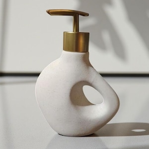 May include: A white ceramic soap dispenser with a gold-coloured pump and collar. The dispenser has an organic shape with a circular opening and a speckled texture. The design is minimalist and modern, suitable for a bathroom or kitchen.