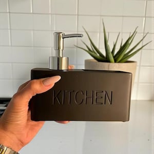 Peut inclure: Un distributeur de savon noir avec une pompe argentée et le mot "KITCHEN" en relief sur le devant. Le distributeur est tenu dans une main, avec un fond carrelé blanc et une plante en pot visible en arrière-plan.