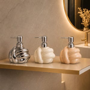 May include: Three soap dispensers with a knotted, rounded shape and a metallic pump mechanism. The first dispenser has a reflective silver finish, the second is a matte cream colour, and the third is a soft peach tone, all arranged in a row.