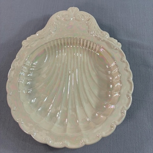 May include: A shell-shaped dish with an iridescent, pearlescent finish. The ceramic piece features a scalloped edge and a central fluted design, with decorative scrollwork at the top. The surface reflects light with subtle rainbow-like tones.