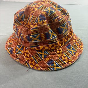 May include: A patterned bucket hat in shades of brown, yellow, blue, and red. The hat features a geometric design and a wide brim. This hat is a stylish accessory.