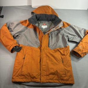 May include: A hooded Columbia jacket in orange and gray. The jacket has a full-length zipper, two front pockets, and a black lining around the hood. The Columbia logo is visible on the chest. The jacket is designed for outdoor activities.