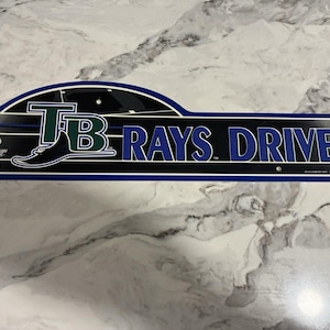 May include: A rectangular sign with the text "TB RAYS DRIVE" in blue and white lettering. The sign has a black background with a blue border and a stylized image of a stingray. The letters "TB" are in green.