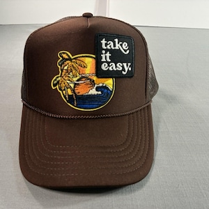 May include: Brown trucker hat with a mesh back and a curved brim. The front features a patch with a tropical scene of a palm tree, sun, and waves, along with the text "take it easy." The hat has a brown rope detail along the brim.