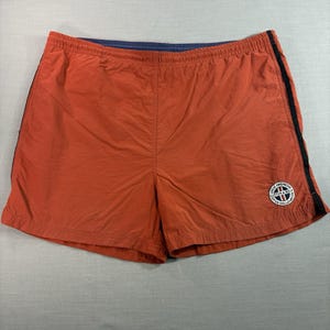May include: Burnt orange athletic shorts with an elastic waistband and a dark blue interior lining. The shorts feature a thin black vertical stripe down each side and a circular white patch on the lower right leg with the text Great American Cutter & Buck.