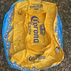 May include: Yellow and blue inflatable pool float with the Corona Extra logo. The float has a rectangular center and a circular outer ring. Text on the float reads "Swim Up Party".