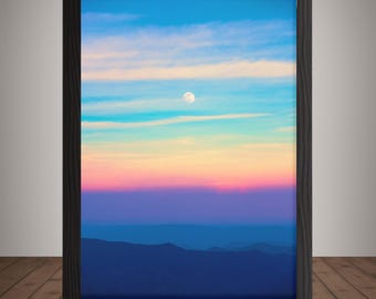 May include: A framed landscape print featuring a vibrant sunset over a mountain range. The sky transitions from blue to pink and purple, with a full moon visible. The frame is black, and the print is displayed on a wooden surface.