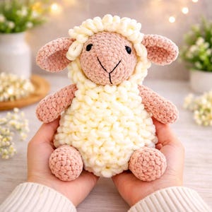 Puede incluir: Un juguete de oveja de ganchillo hecho a mano en colores pastel suaves. La oveja tiene un cuerpo color crema, una cara, orejas y extremidades de color rosa claro, y ojos bordados en negro. El juguete está diseñado para ser tierno y se sostiene en las manos.