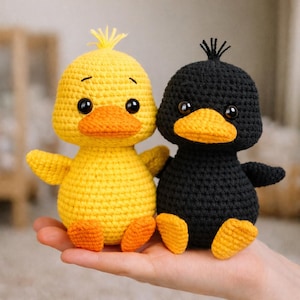 Op de afbeelding: Twee gehaakte eendjes, een gele en een zwarte, worden getoond. Het gele eendje heeft een oranje snavel en poten, met een kleine pluk geel garen op zijn hoofd. Het zwarte eendje heeft een oranje snavel en poten, met een kleine pluk zwart garen op zijn hoofd.