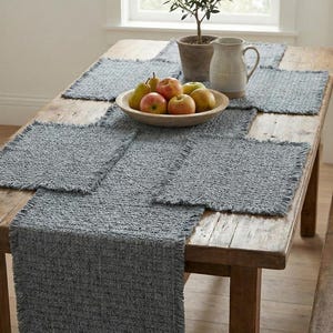 Puede incluir: Una mesa de madera rústica está decorada con un camino de mesa tejido gris y manteles individuales a juego. Un cuenco de madera contiene manzanas y peras. Una pequeña planta en maceta y una jarra crema también están sobre la mesa.