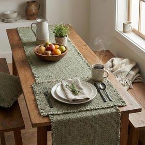以下が含まれることがあります： 食事のためにセットされた素朴なダイニングテーブル。テーブルには、緑色のテクスチャードランナーとプレースマットが飾られています。木製のフルーツボウル、ピッチャー、コーヒーマグがあります。白いお皿、カトラリー、ナプキンがセッティングを完成させています。
