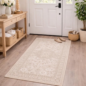 Op de afbeelding: Beige rechthoekig vloerkleed met bloemenpatroon, afmeting 69 cm x 114 cm. Het vloerkleed ligt op een houten vloer voor een witte deur. Het kleed heeft een decoratieve rand en een centraal medaillonontwerp.