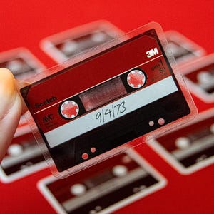 Puede incluir: Una pegatina transparente de una cinta de cassette retro con detalles rojos y negros. La cinta tiene el texto "Scotch" y "9/4/73" escrito en ella. La pegatina se sostiene sobre un fondo rojo, con otras pegatinas similares visibles.